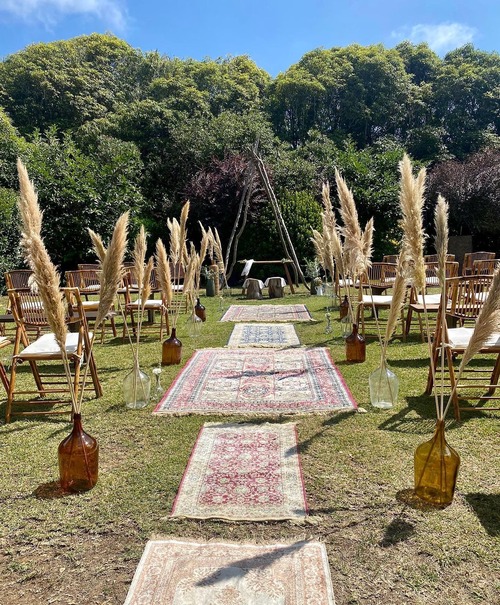 Casamento ao ar livre com tapetes vintage a formar o corredor até um altar rústico de madeira. Garrafões de vidro com erva-dos-pampas decoram o caminho, e cadeiras de madeira com almofadas brancas estão dispostas para os convidados