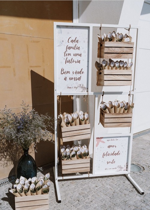 Estrutura branca com placas decorativas e caixas de madeira penduradas, contendo lembranças para os convidados. As placas têm mensagens de boas-vindas. Ao lado, um vaso de vidro escuro com ramos secos complementa a decoração ao ar livre.