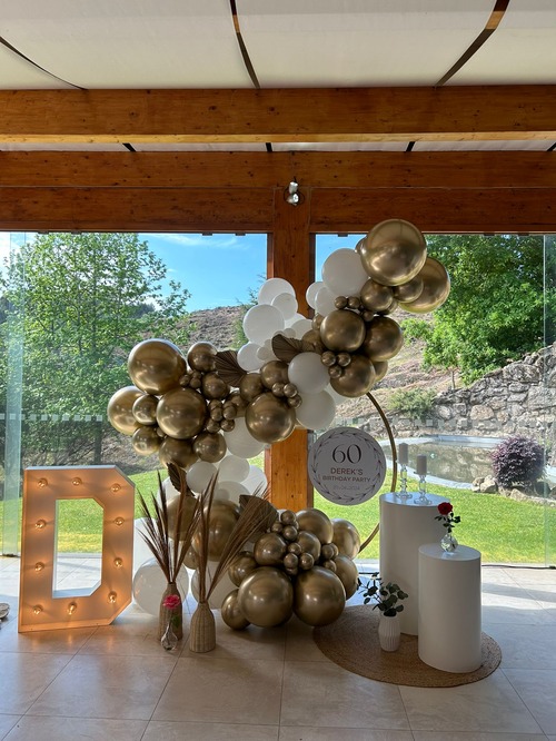 Decoração de festa de 60 anos com balões dourados e brancos, um grande 'D' iluminado e um letreiro em um ambiente ao ar livre com vegetação ao fundo.
