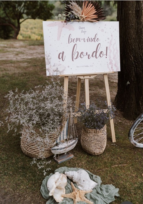 Placa de boas-vindas para um evento com tema náutico, com
                uma placa de madeira escrita 'Bem-vindo a Bordo!' em um
                cavalete, rodeada por decoração náutica, como conchas, um barco
                à vela em miniatura e flores em cestos de vime.
